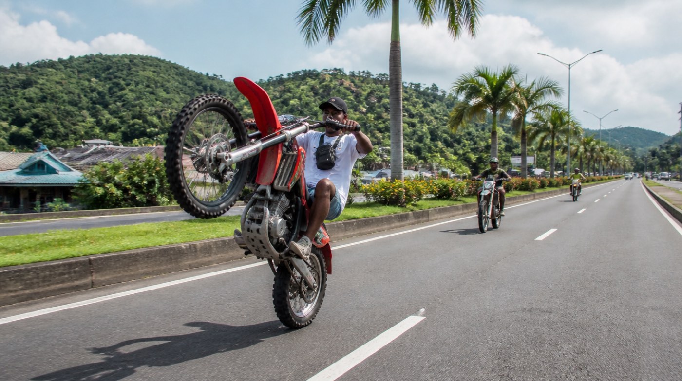 Un pilote lève la roue avant de sa 125 cross rouge sur une voie rapide de Martinique, deux autres suivent, palmiers et mornes verts au bord de la route.